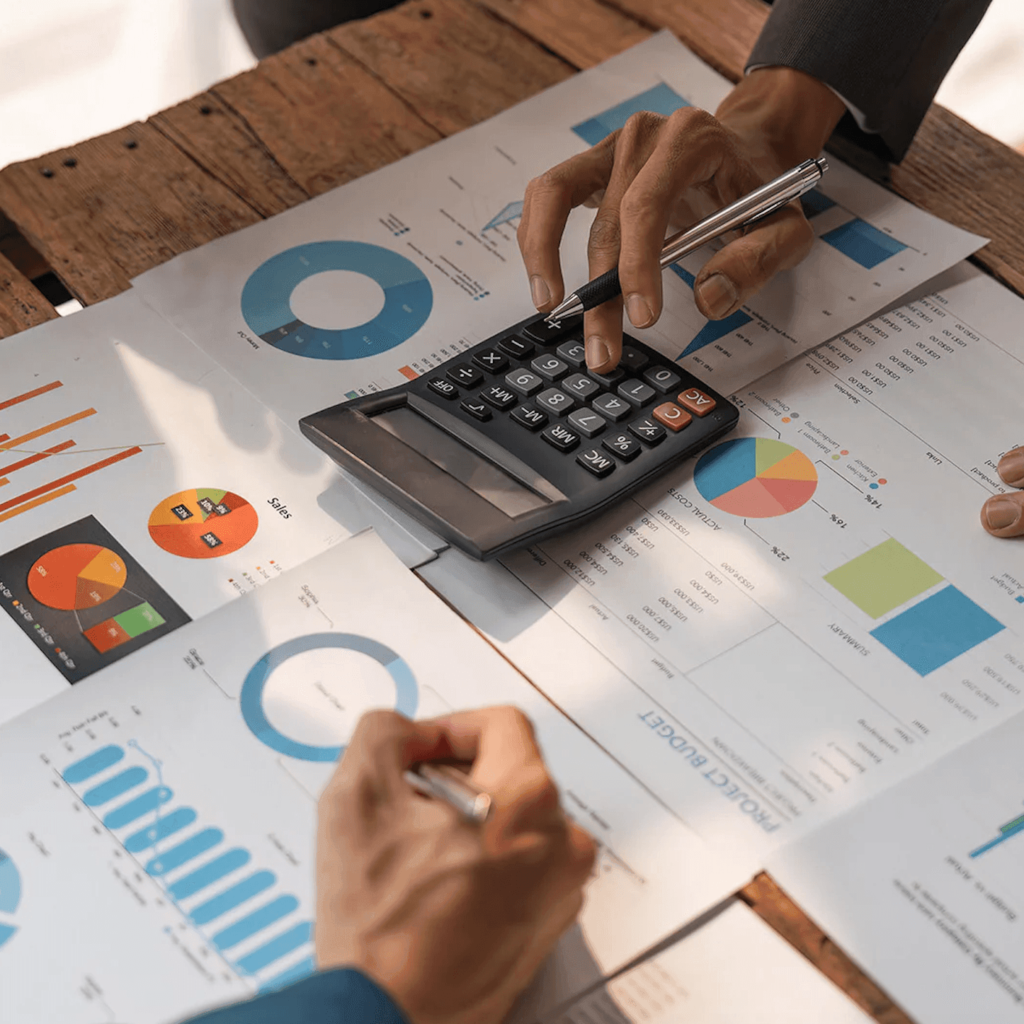 Hands working with a calculator and analyzing financial charts and graphs on a wooden table.