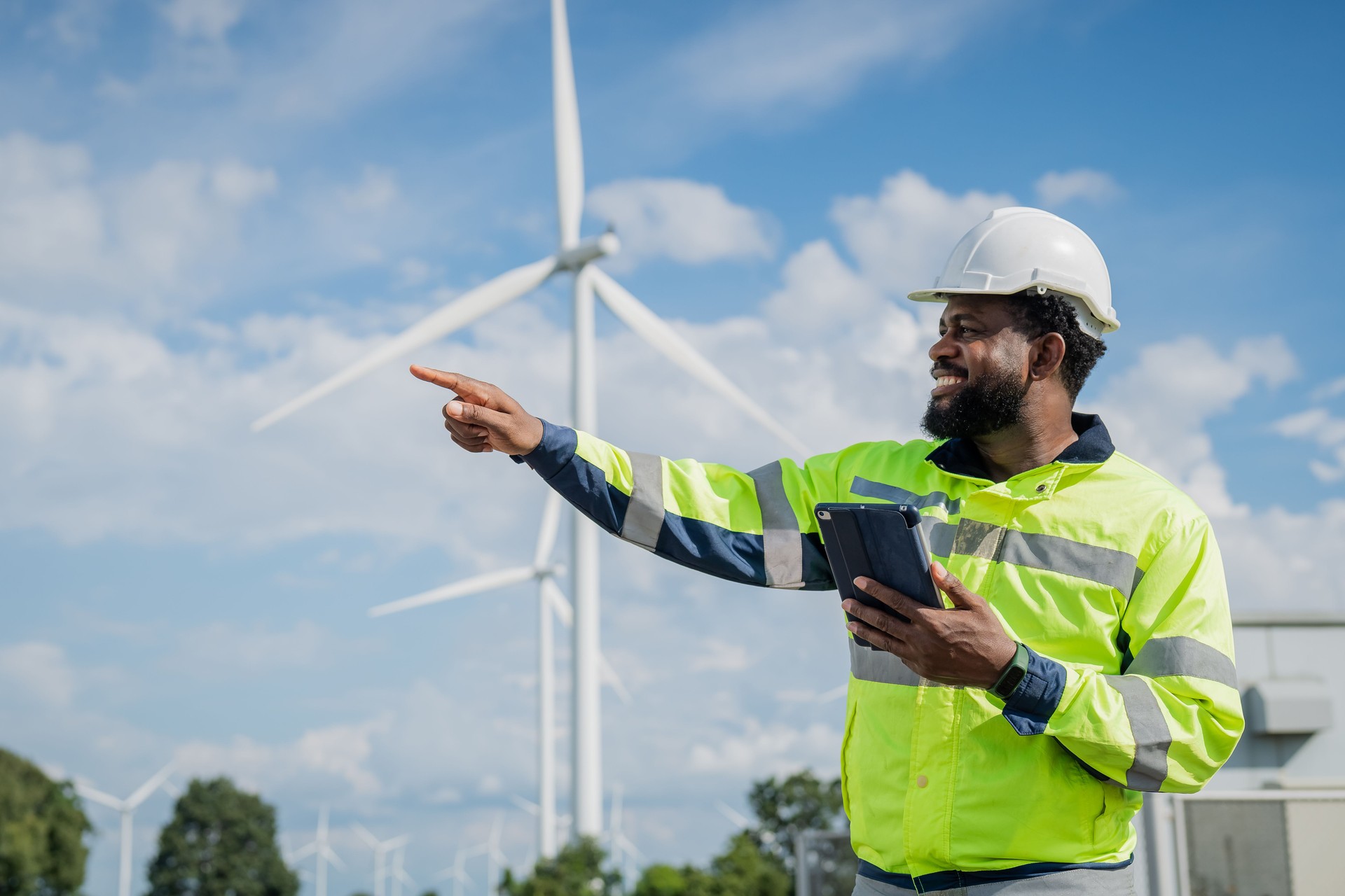 Un hombre con un chaleco de seguridad amarillo y negro señala una turbina eólica. Está sonriendo y está feliz. eléctrico, ingeniero, medio ambiente, energía, limpio, sostenible, turbina eólica, naturaleza